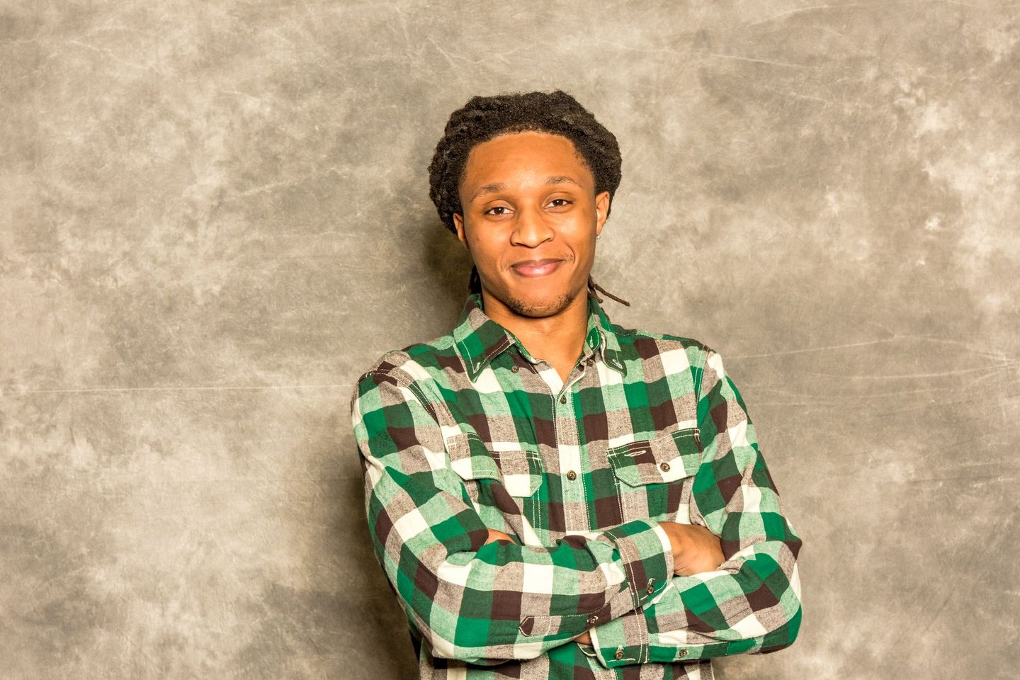 Person in a green-and-white plaid shirt standing with arms crossed against a gray backdrop.