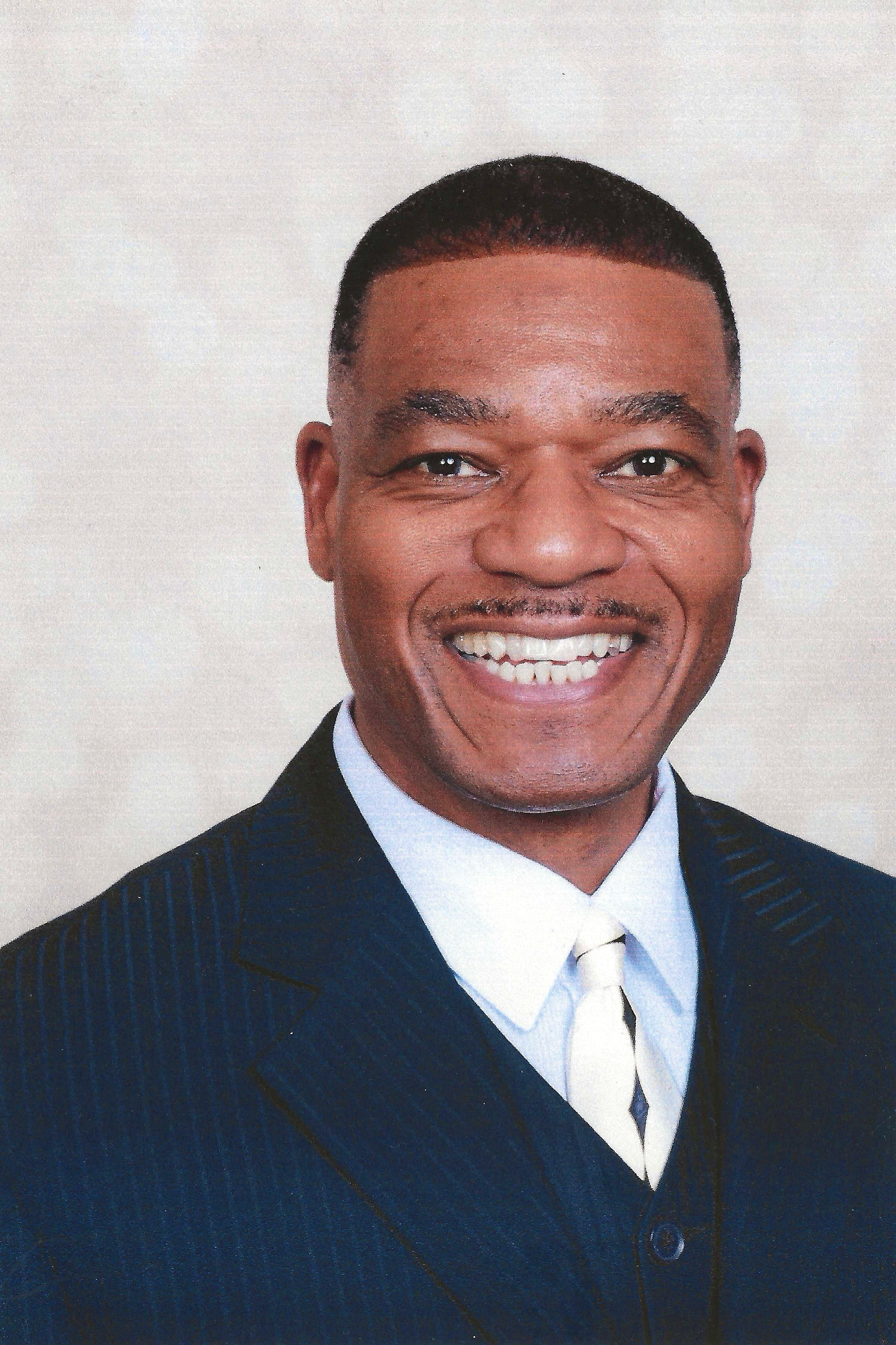 Smiling man in a dark suit and white tie against a plain background