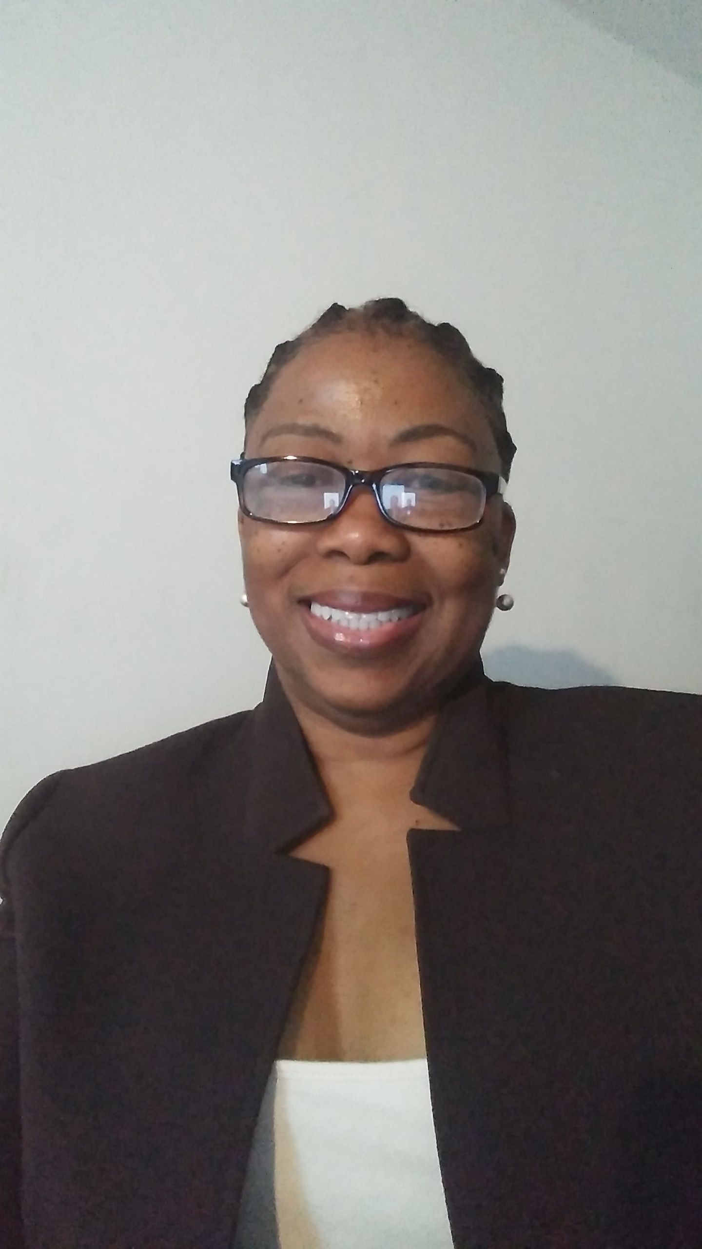 Smiling person in glasses and a dark blazer, standing against a plain wall.
