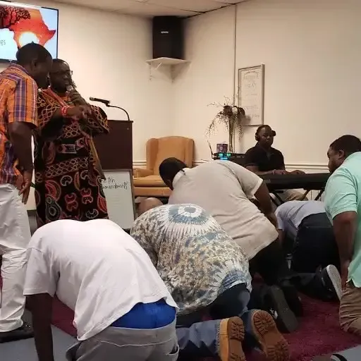 People kneel in prayer in a small room while a speaker stands at a podium.