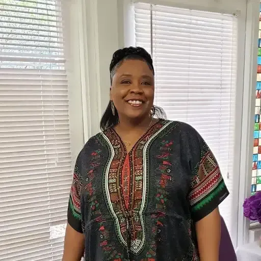 Smiling woman in a patterned black, green, and red top standing in a bright room with white blinds.