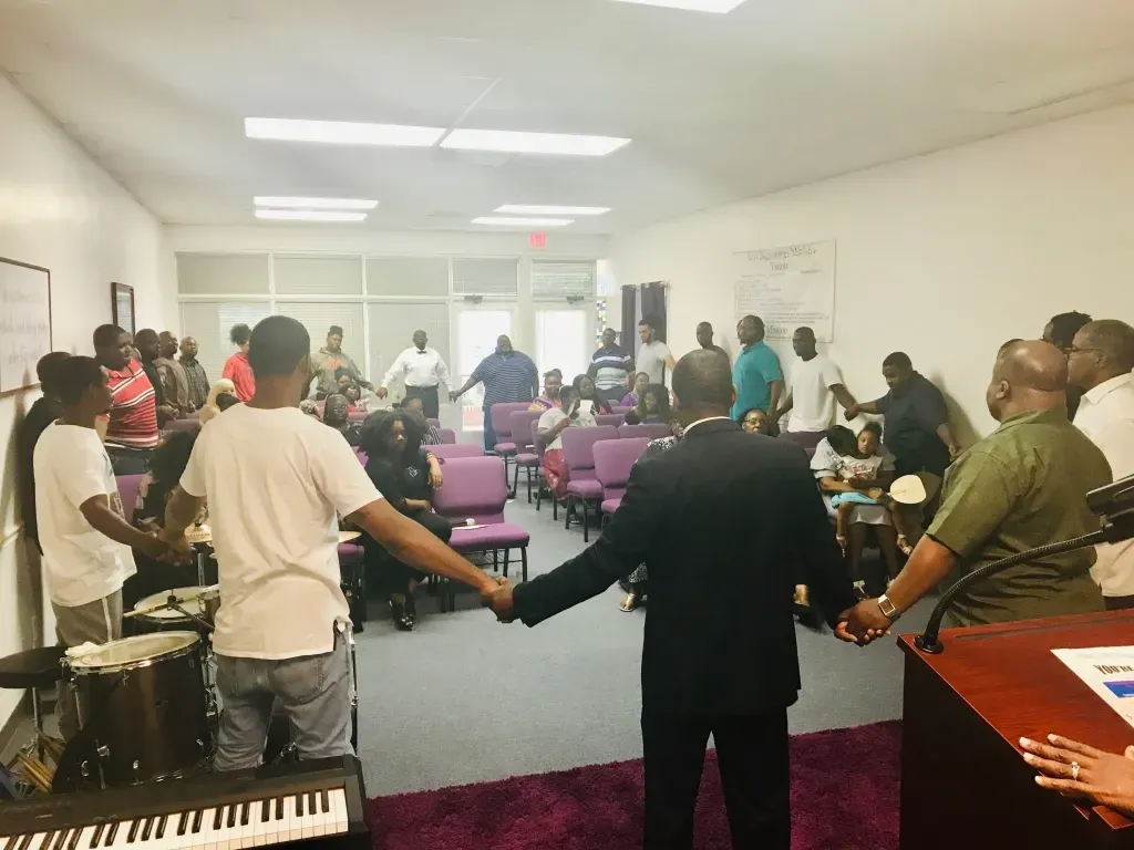 People standing in a circle holding hands in a bright meeting room with purple chairs and a keyboard in front.