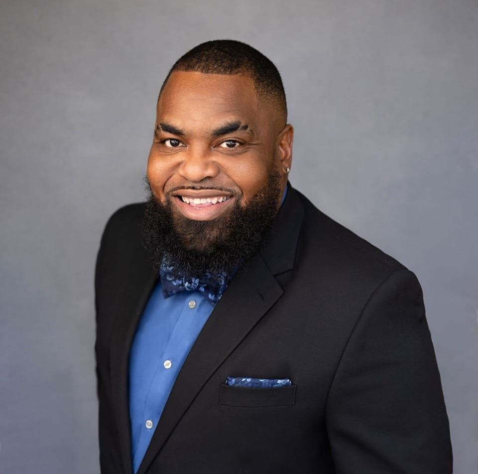 Smiling man in a black suit jacket and blue shirt against a gray background