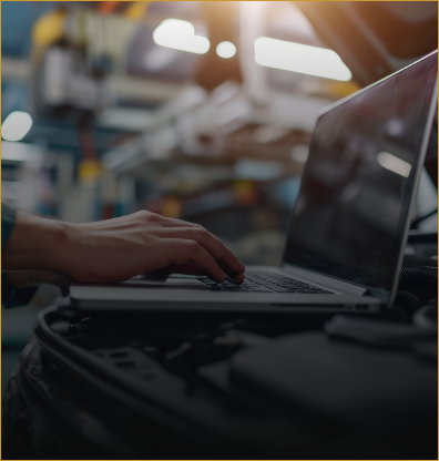 Person typing on a laptop beside a car in a dim garage, with a red light glowing in the background | The Family Mechanics - Cherry Hill