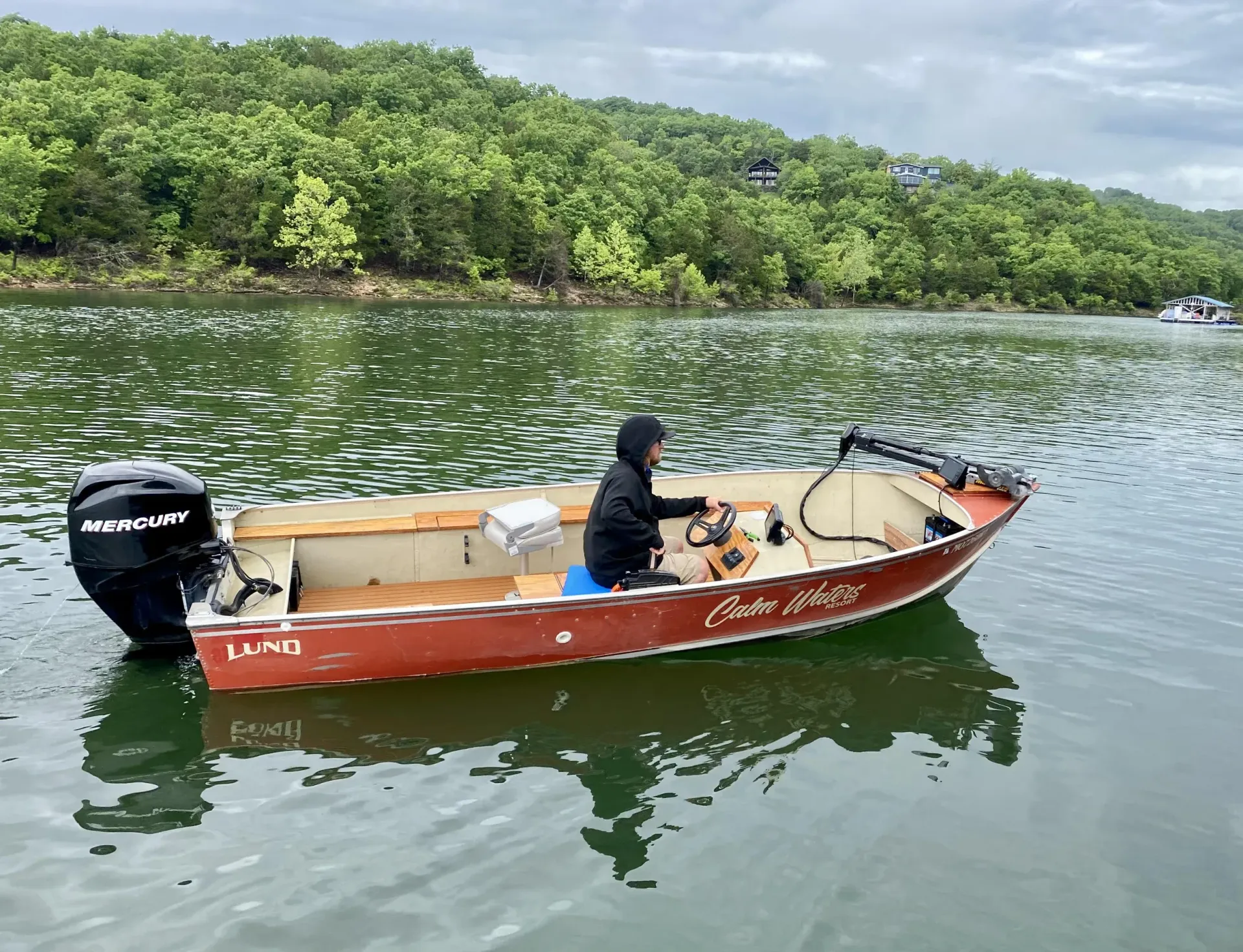 Person piloting a small motorboat on a lake. Hills covered with trees are in the background.