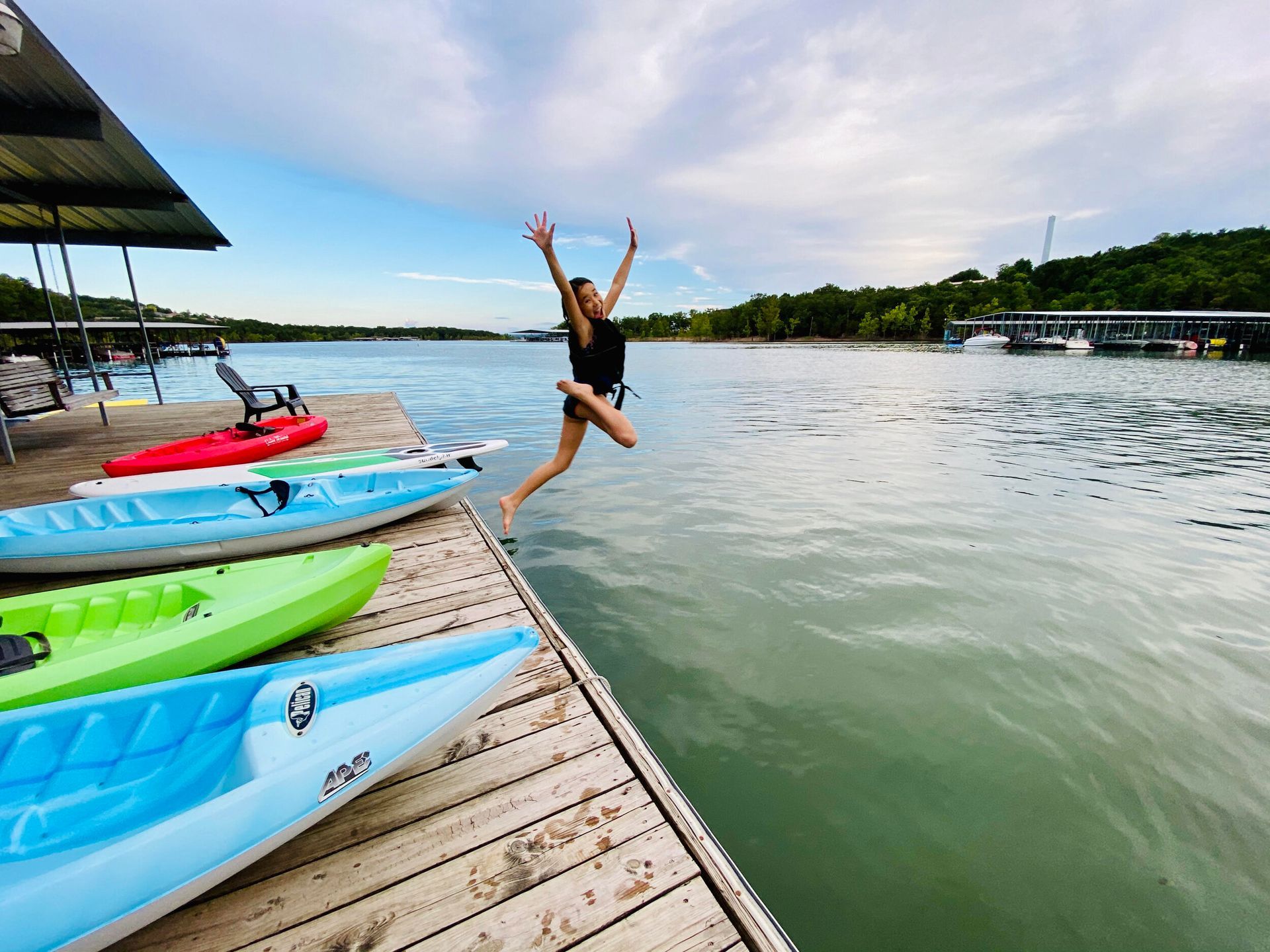 Woman jumps off dock into water, arms raised, kayaks alongside.