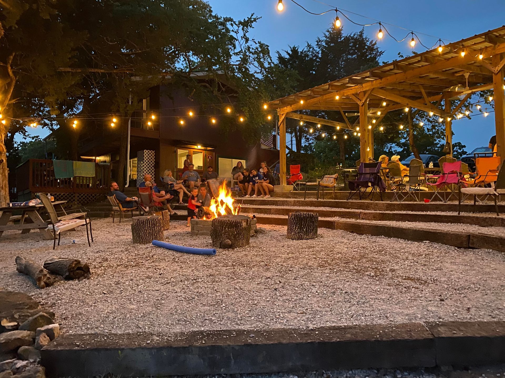 Outdoor gathering around a fire pit with seating. String lights illuminate a wooden structure and people.