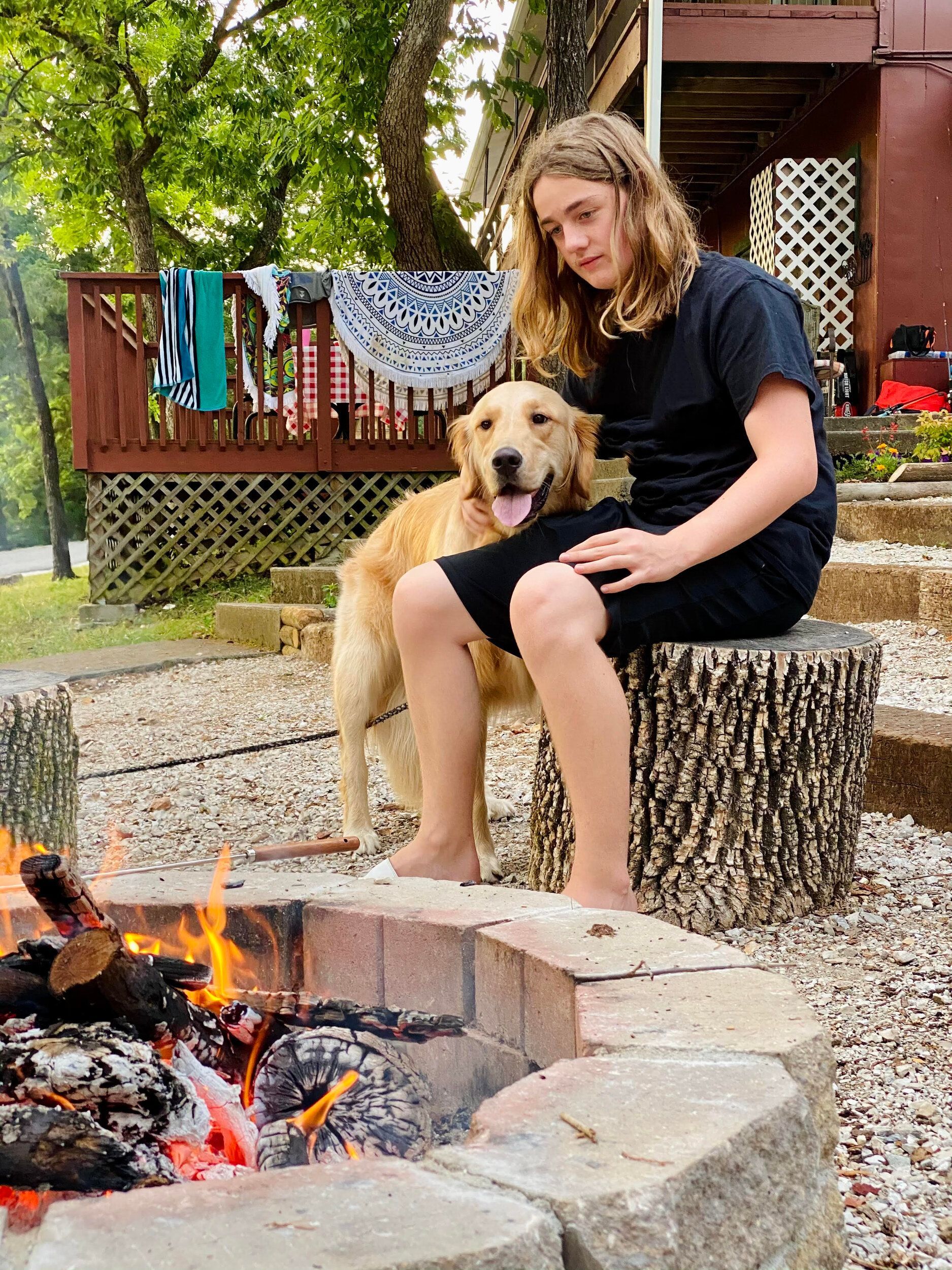 Teen sits with dog near a bonfire; backyard setting.