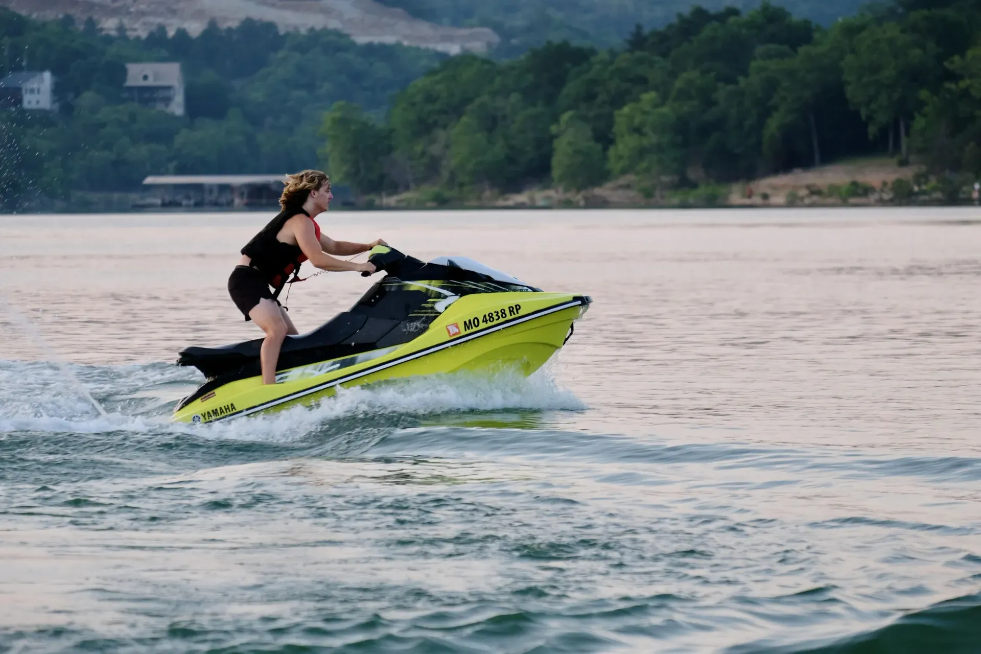 Woman on a yellow jet ski, speeding across water near a forested shoreline.