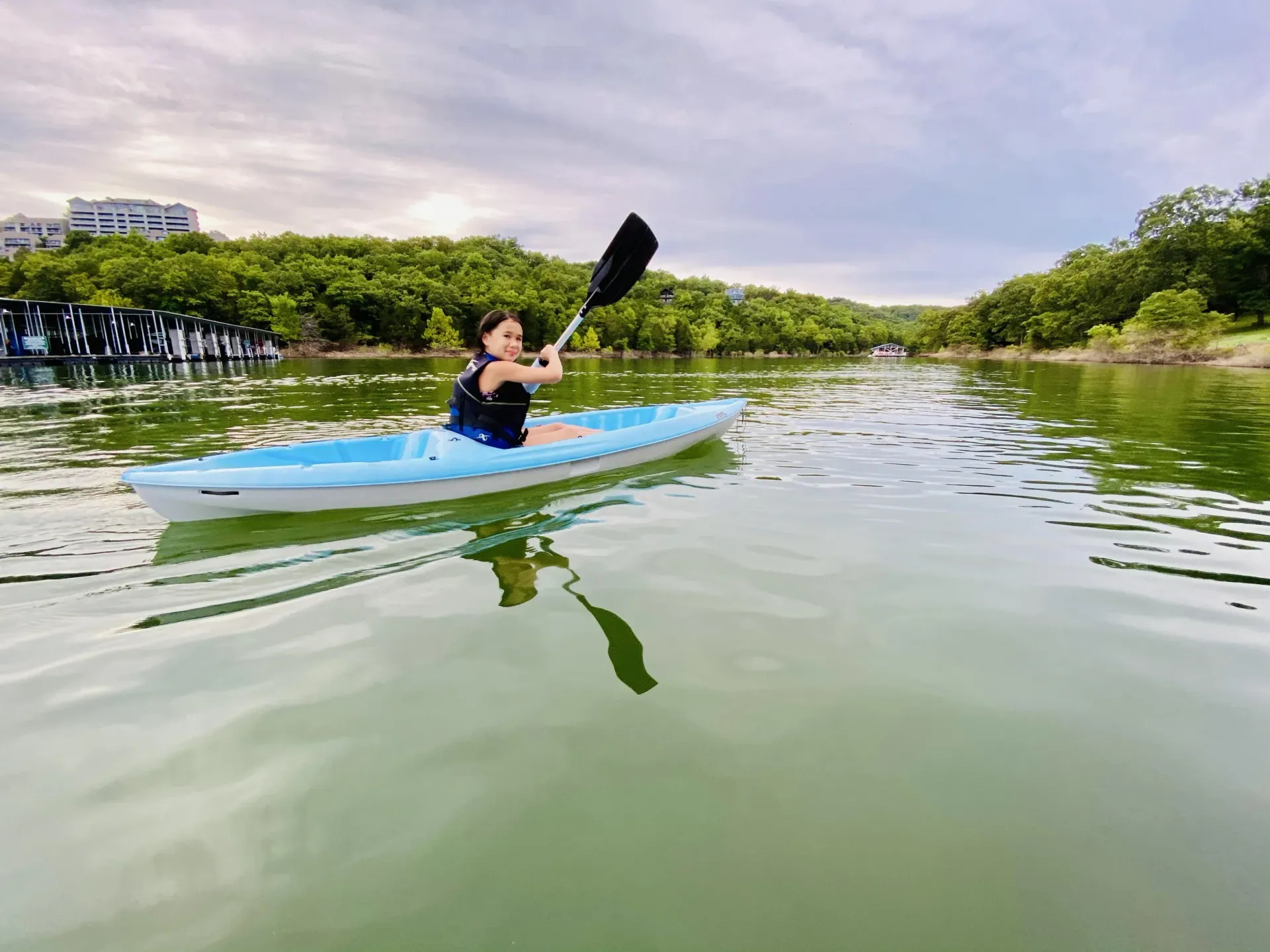 Woman kayaking on a calm lake, surrounded by trees and overcast sky; blue kayak and paddle.