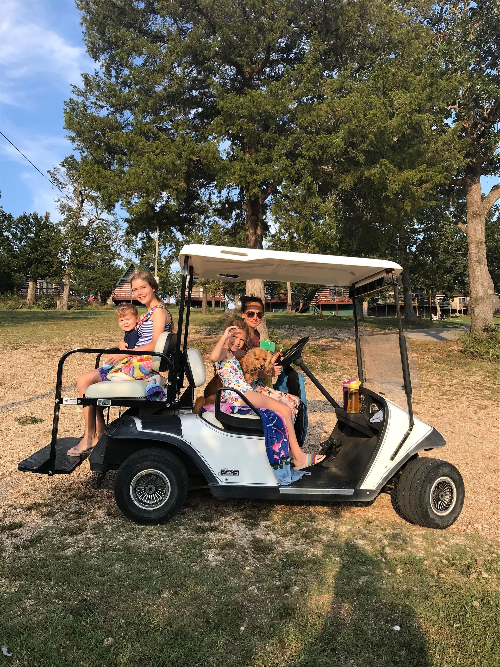 People riding in a white golf cart on a grassy area, trees in the background, blue sky.