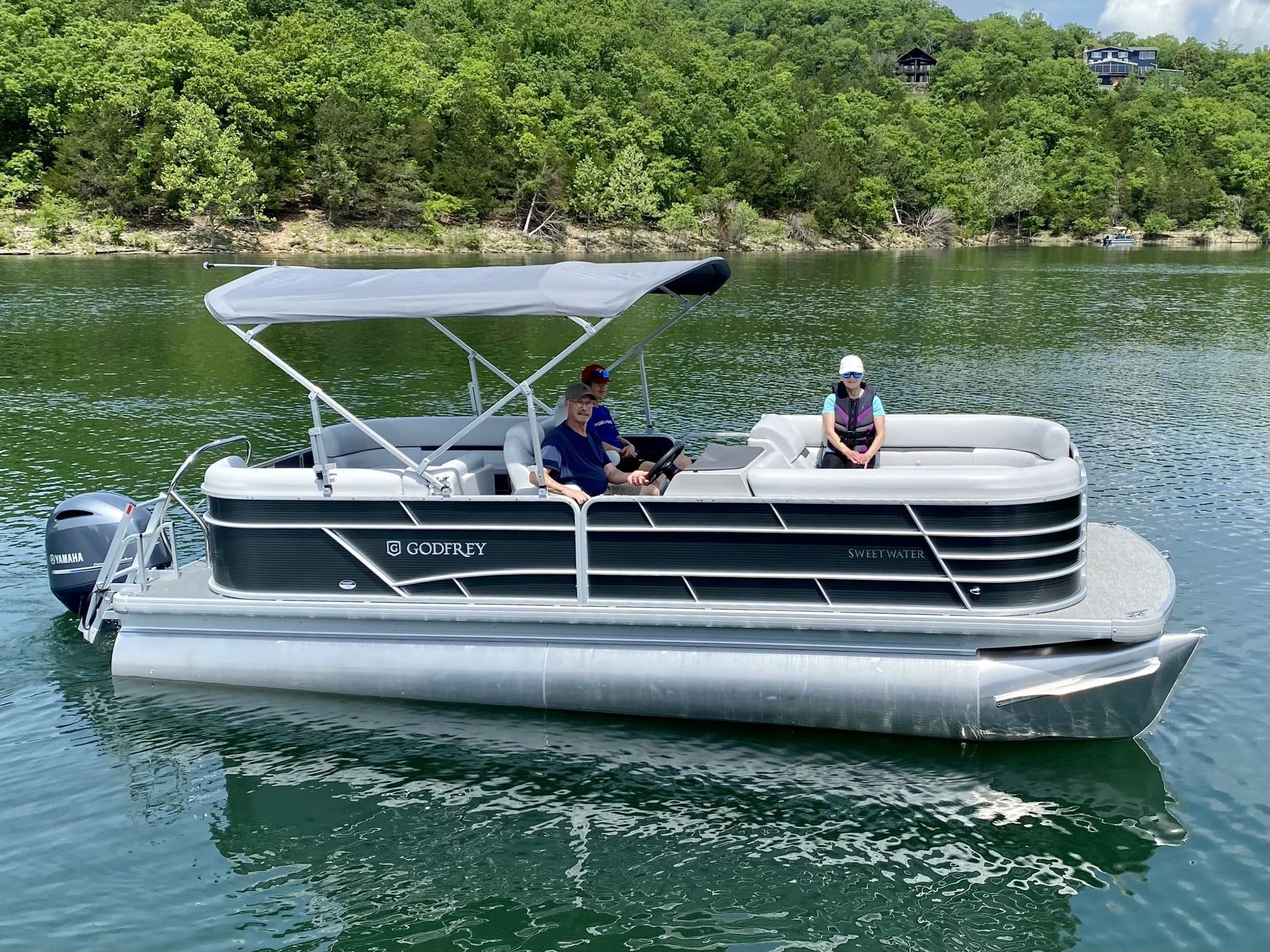 People waving from a pontoon boat on a lake; sunny day. Dog sitting on the front.