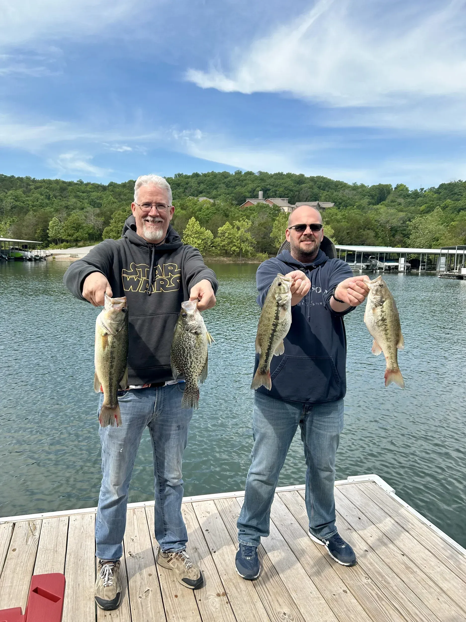 Two men holding up fish on a dock at a lake. Sunny day with trees and blue sky.