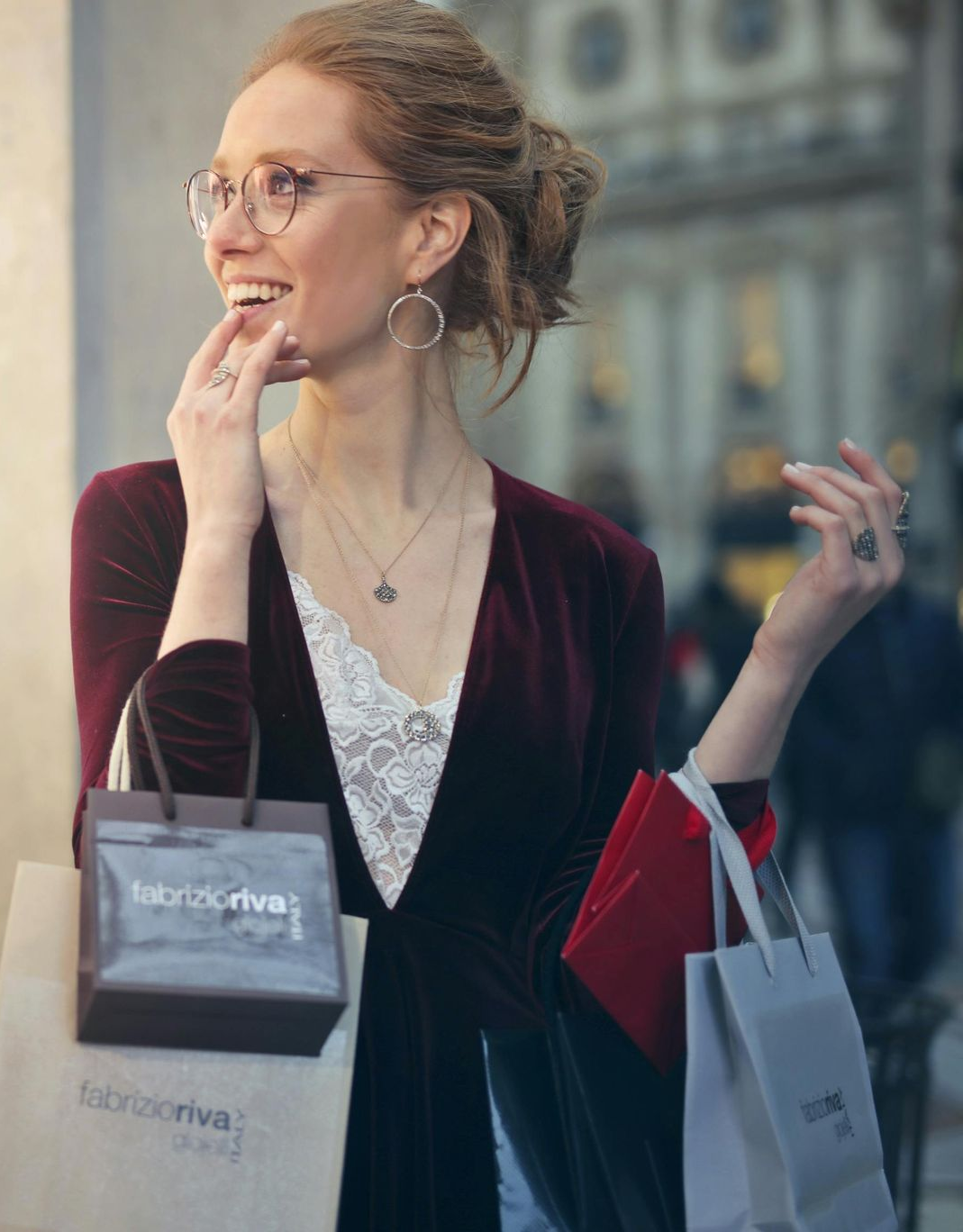 Woman smiles, holding shopping bags, in front of a building. She wears glasses, jewelry, and a velvet jacket.