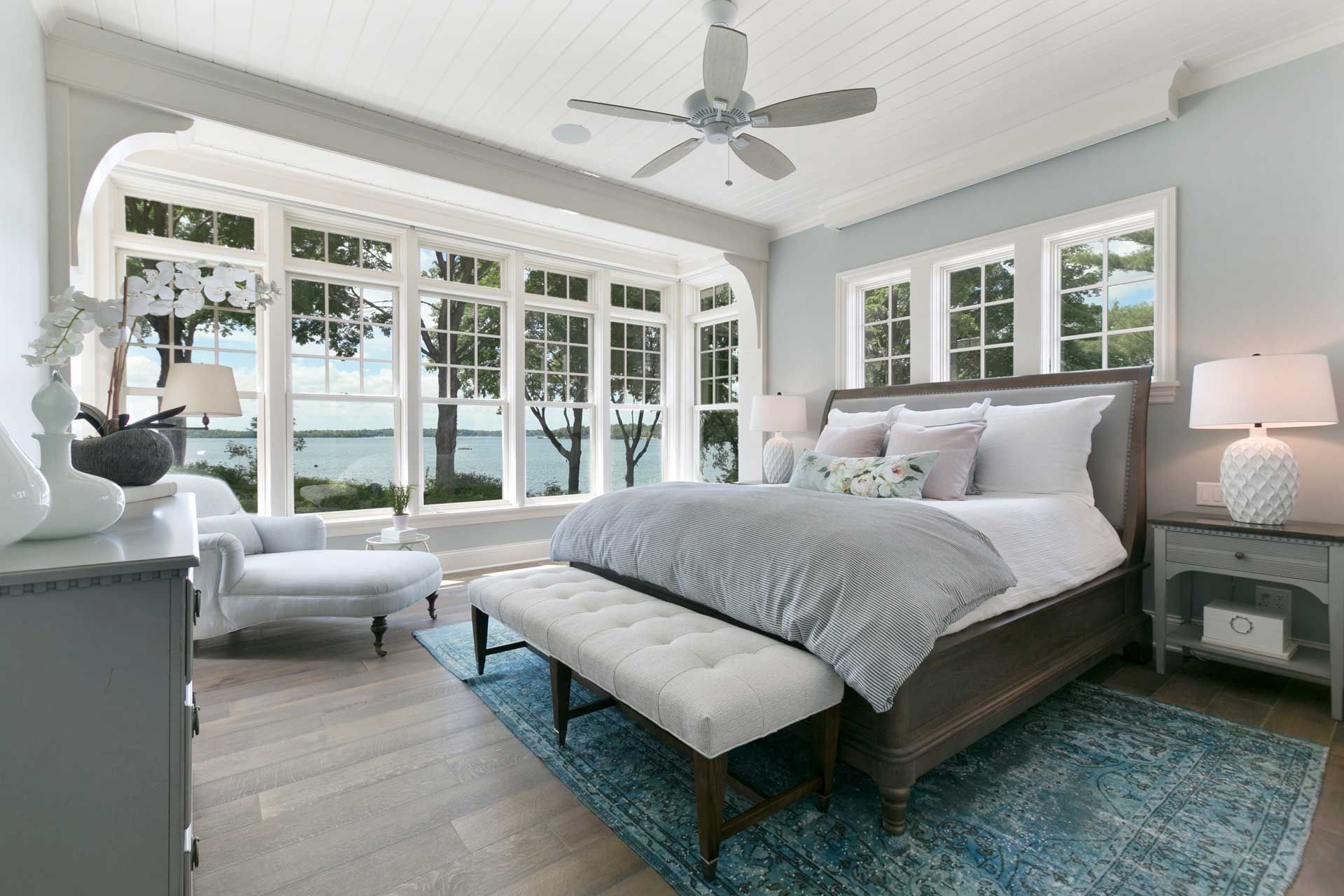 A bedroom with a large bed , a bench , a ceiling fan and lots of windows.
