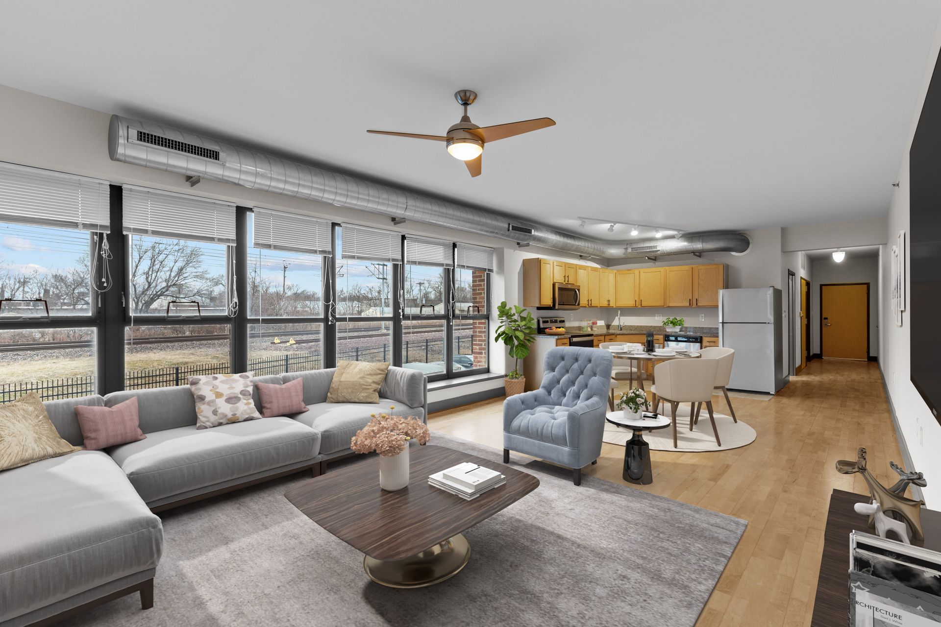 Bright open-concept living room with sectional sofa, coffee table, dining area, and kitchen by large windows.