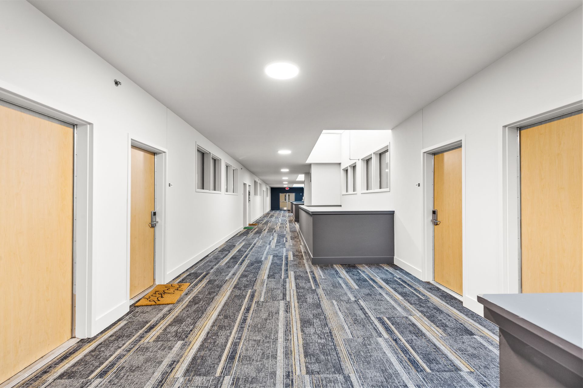 Bright, empty hallway with white walls, wood doors, gray carpet, and a reception counter