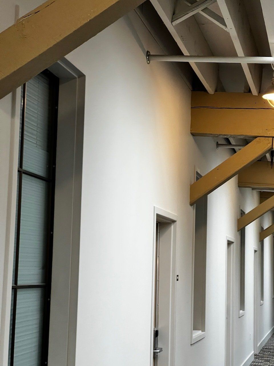 Covered outdoor corridor with white walls, windows, and yellow support beams under a slanted roof