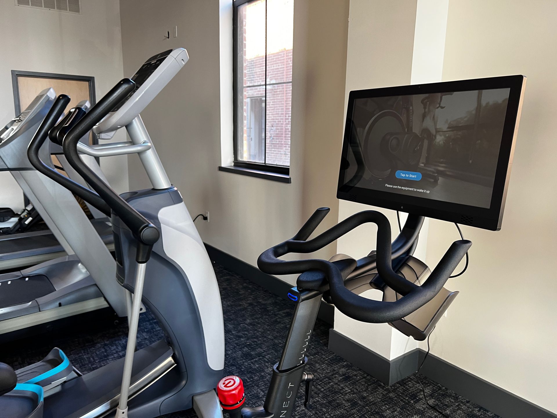 Gym with elliptical machine and stationary bike facing a wall-mounted TV near a window.