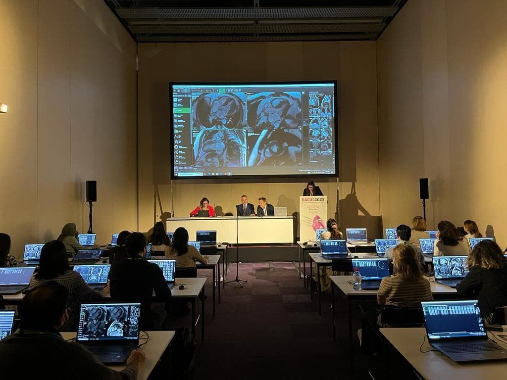 Conference room with attendees at laptops facing a screen showing medical images; speakers on stage.