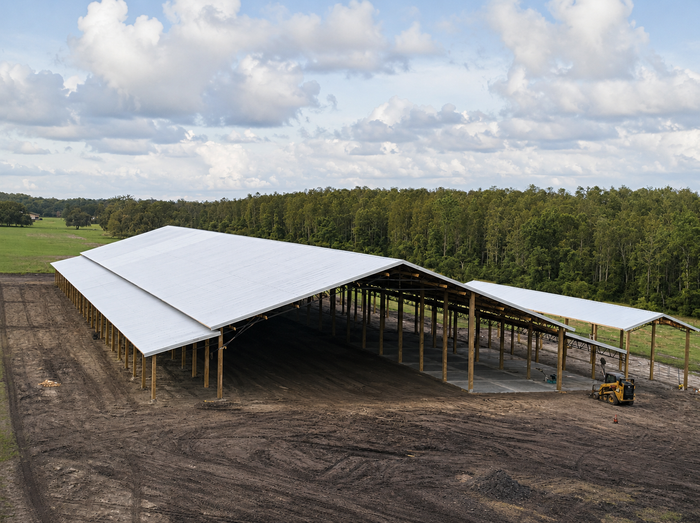 Large white-roofed farm structure under construction on a dirt site, with a small loader nearby