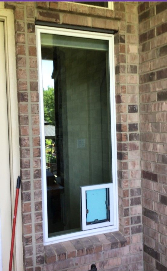 A window with a dog door in it is on a brick wall.