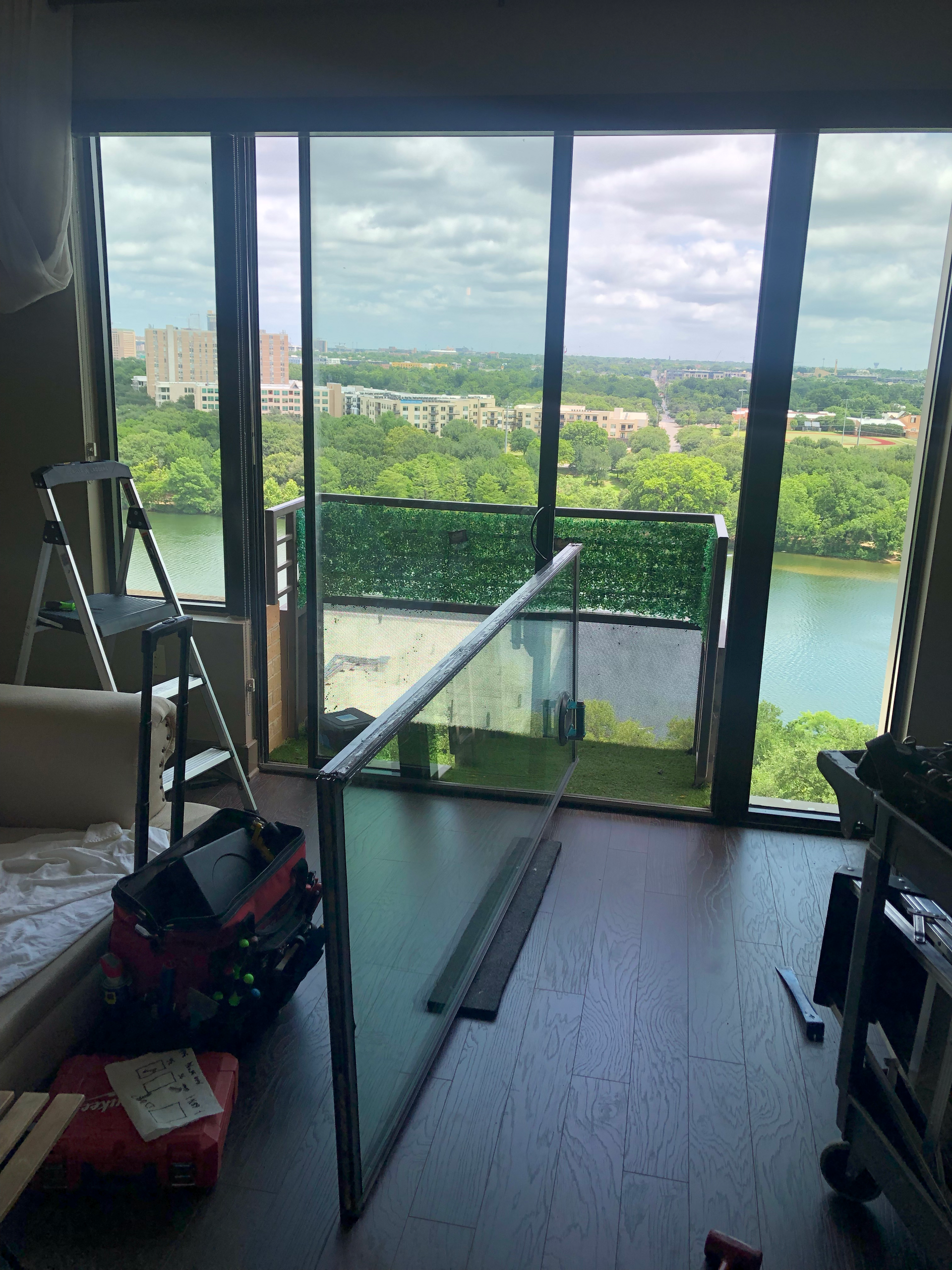 Interior view of a room with a sliding glass door open to a balcony with green foliage and a city view. Tools are visible.