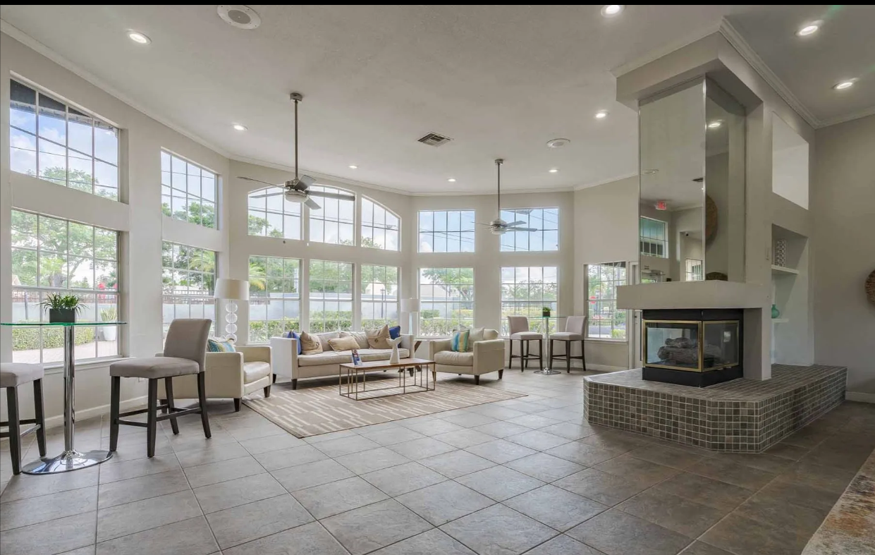 Bright, spacious resident clubhouse lounge area with large windows, seating area, fireplace, bar stools, and tiled floor. Discover the lifestyle waiting in our Clearwater, FL apartments for rent—book today.