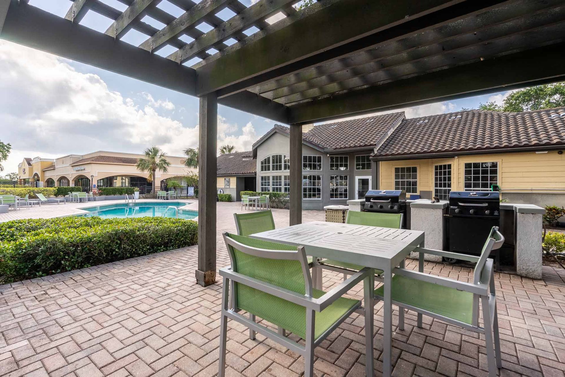 Patio with table, chairs, grills, near a pool, under a wooden pergola on a brick paved surface. See the layout in person with a tour of our 2-bedroom apartments in Clearwater, FL.