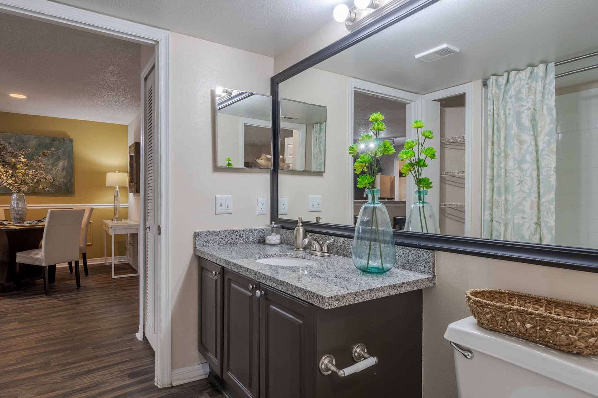Bathroom with dark vanity, granite countertop, large mirror, and open doorway to dining area. Visit our Clearwater, FL apartments for rent and picture your routine here.