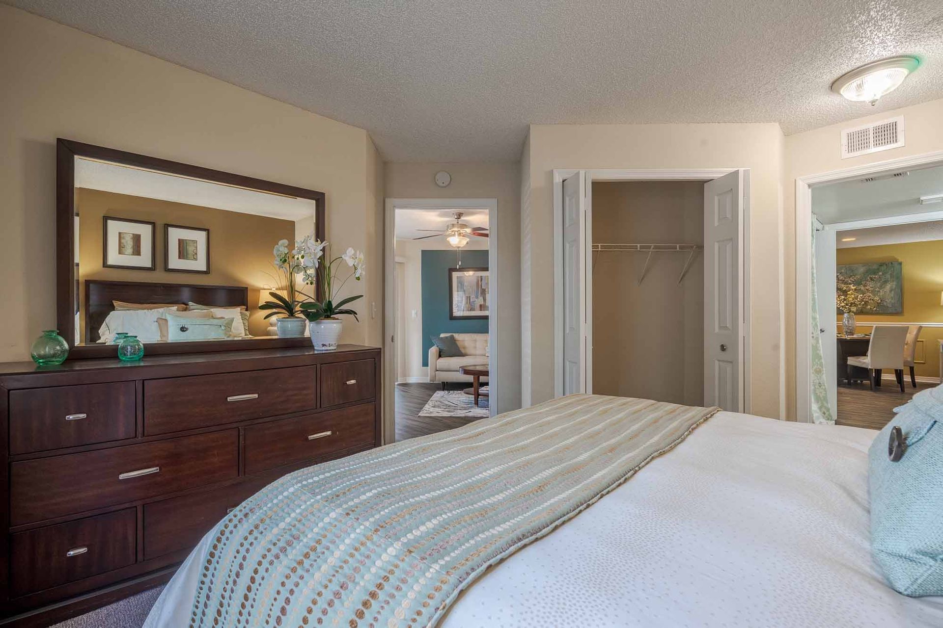 Bedroom with a dark wooden dresser, large mirror, bed, and closet; a glimpse into a living area. Schedule a private tour of our Apartments for Rent in Clearwater, FL today.