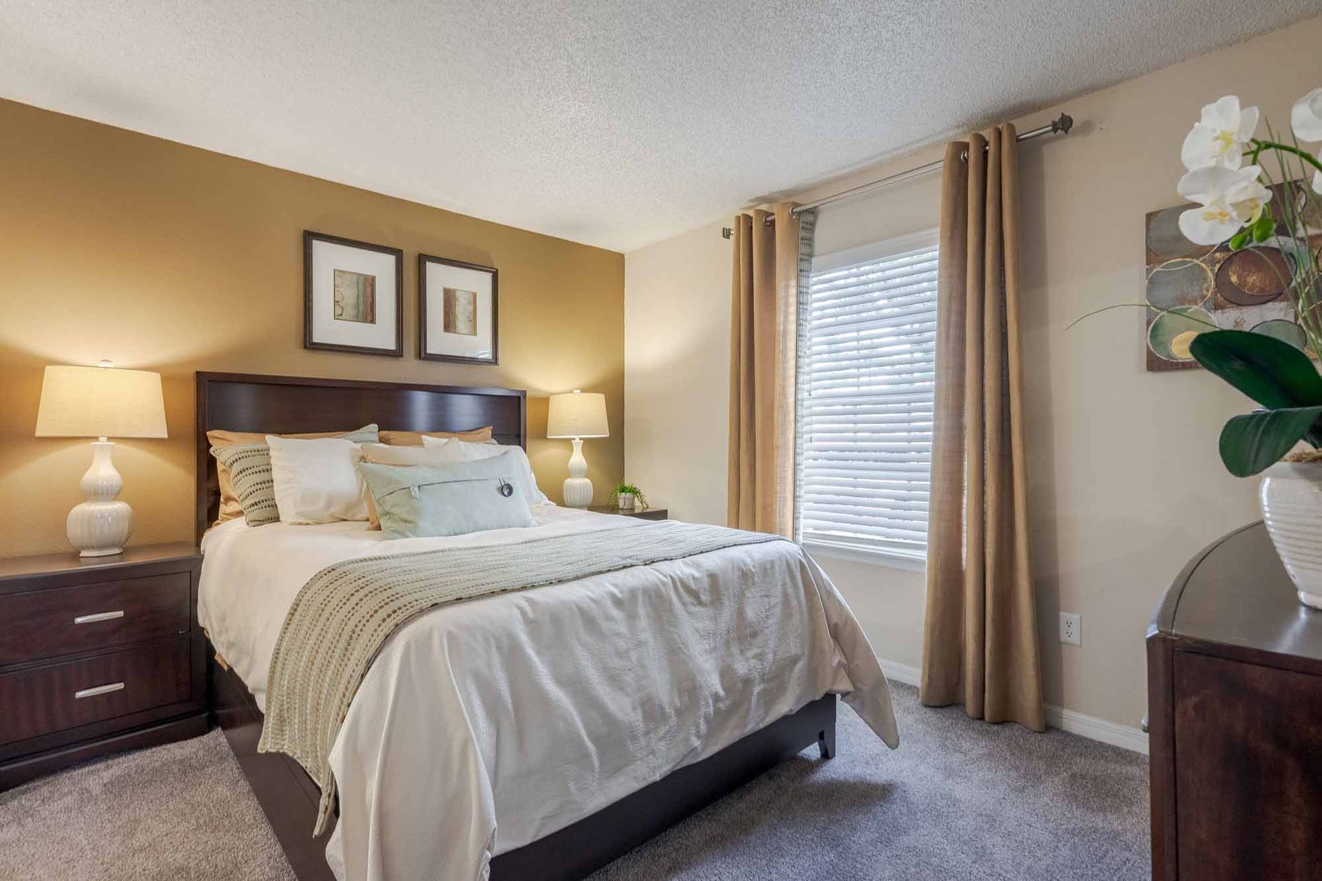 Bedroom with a bed, nightstands, lamps, window with curtains, and artwork. Warm color palette. Explore our expansive 3-bedroom apartments in Clearwater, FL with an in-person visit.