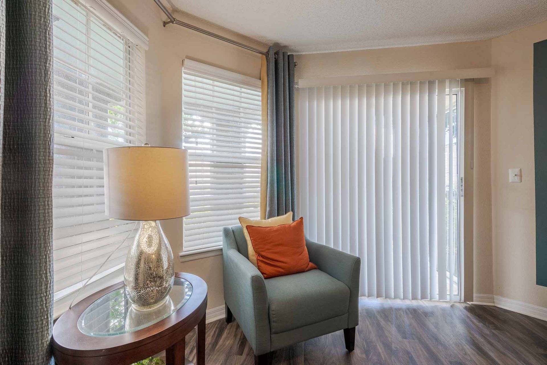 Cozy corner with armchair, lamp, and window, curtains partially open, showing blinds, orange pillow, and sliding glass door. Tour our spacious 2-bedroom apartments in Clearwater, FL and find your fit.