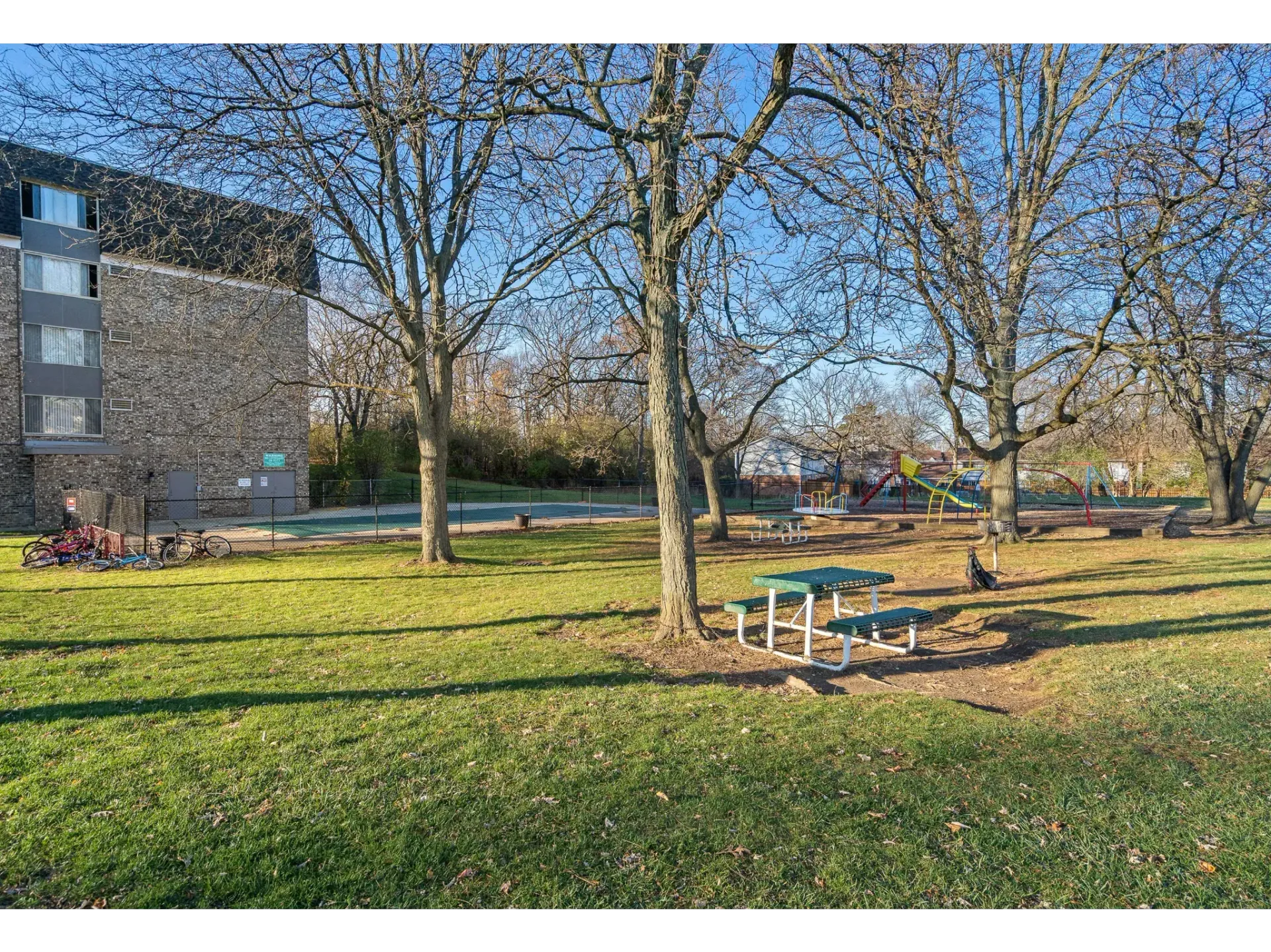 View of a community park with trees, picnic tables, playground, and a building in the background.