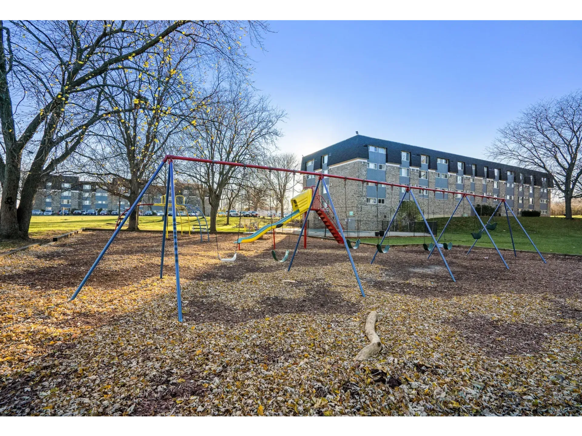 Playground area with swings and slide surrounded by autumn leaves and an apartment building in the background.
