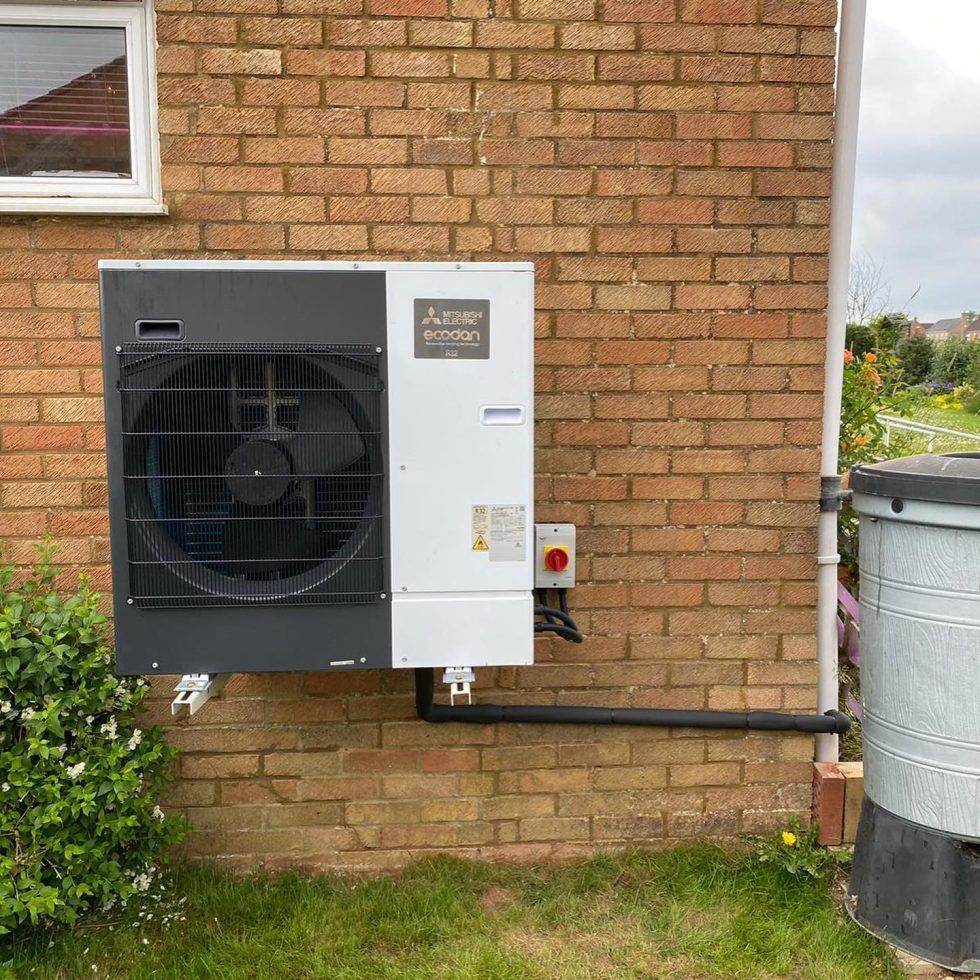 A Mitsubishi Ecodan heat pump unit mounted on a brick exterior wall next to a vertical pipe and a grey container.