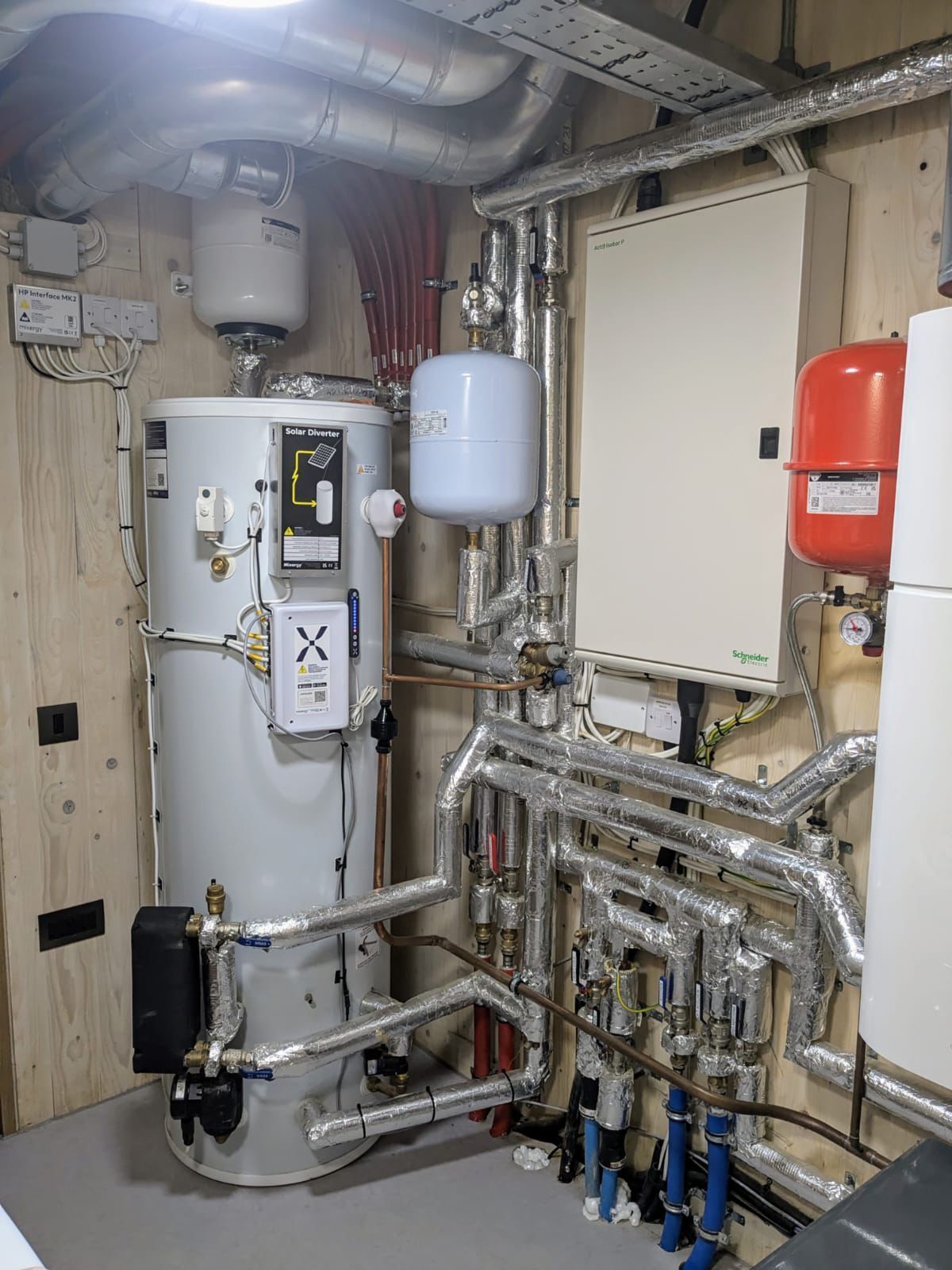 Mechanical room featuring a white cylindrical water heater, various insulated pipes, and red and white expansion tanks.