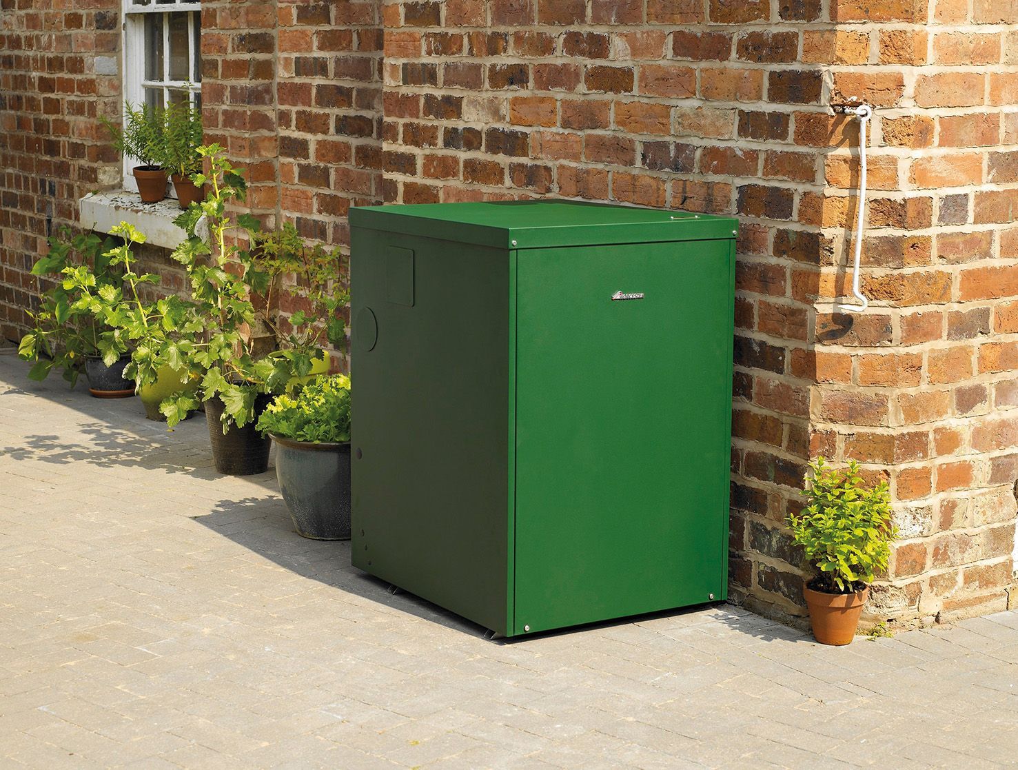 A green metal outdoor storage cabinet sits on a patio against a red brick wall, surrounded by several potted plants.