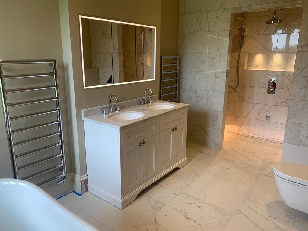 A modern, neutral-toned bathroom featuring a double vanity, a lit mirror, a walk-in tiled shower, and a heated towel rack.