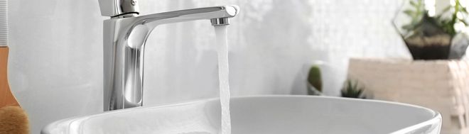 A chrome bathroom faucet flows with water into a white sink, with a small decorative plant in the background.