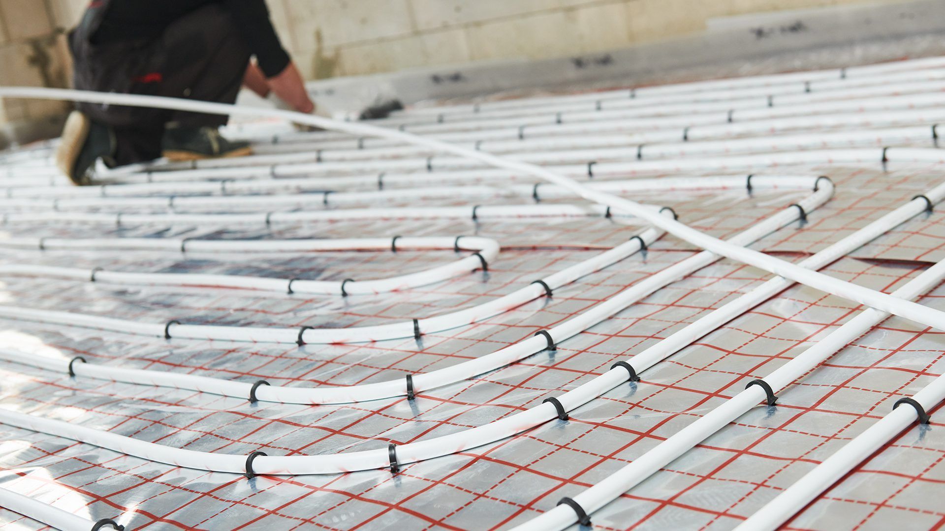 A worker installs white tubing for a radiant floor heating system over reflective foil insulation on a grid pattern.