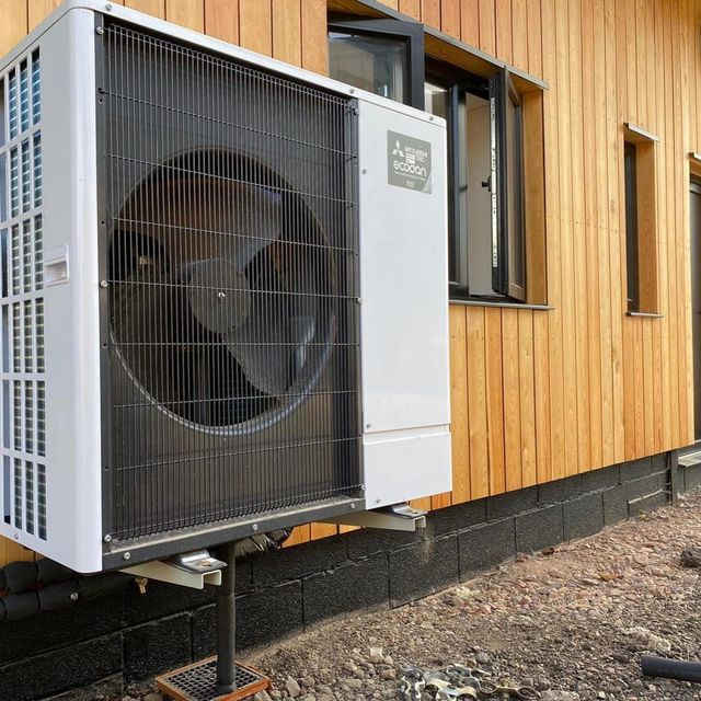 A white Mitsubishi heat pump unit mounted on a pole outside a building with vertical wooden siding.