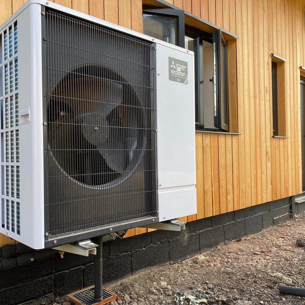A Mitsubishi Ecodan heat pump unit installed outdoors on a metal stand next to a wooden-sided building.