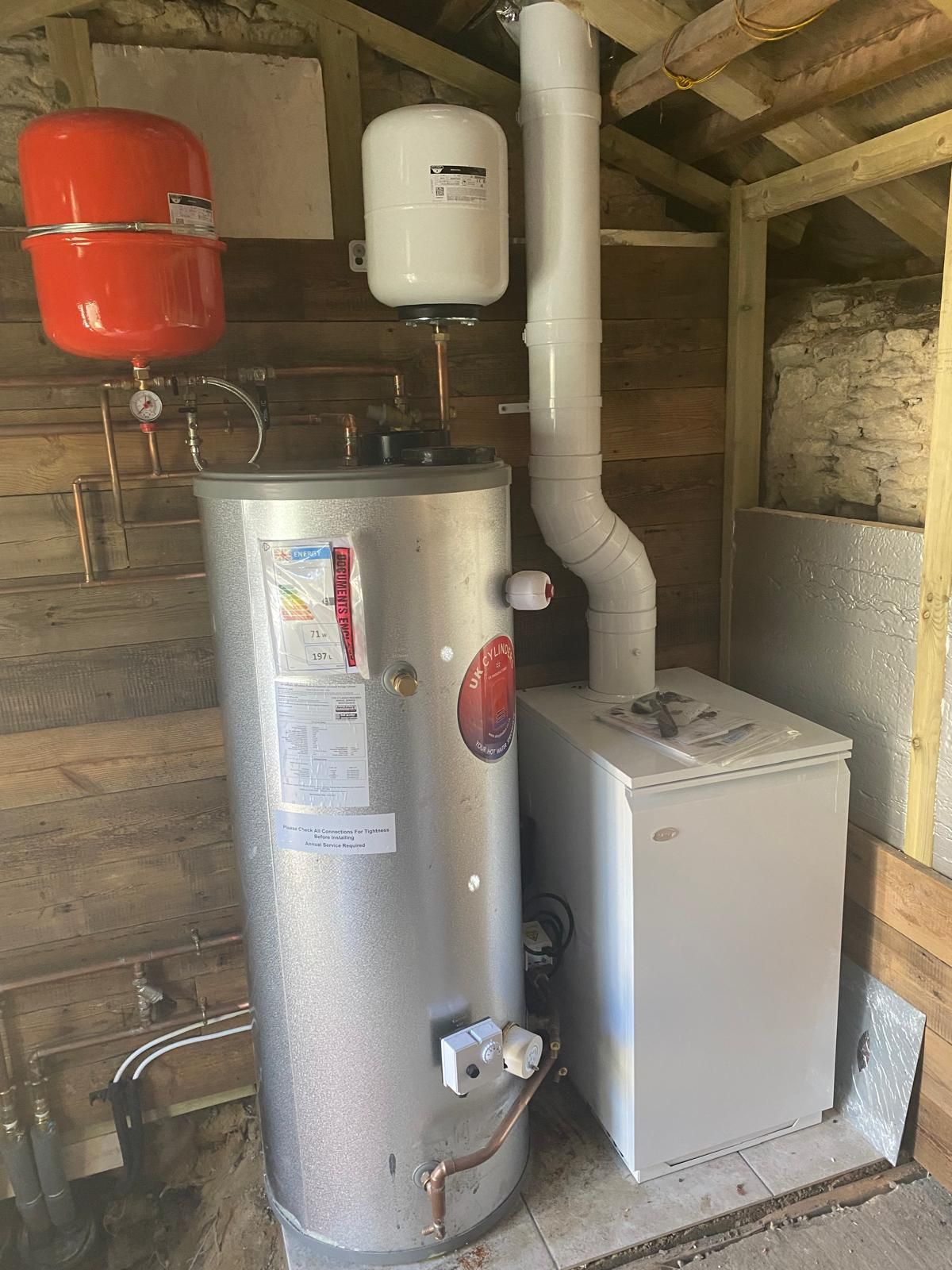 A metal hot water cylinder, red and white expansion vessels, and a white boiler with a flue in a wooden-framed utility room.
