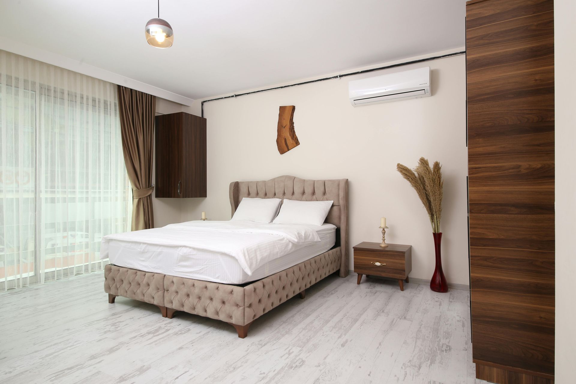 A modern bedroom with a tufted beige bed, wood furniture, neutral decor, and large floor-to-ceiling windows.