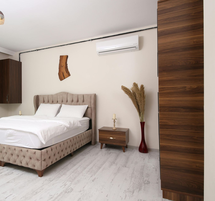 A bedroom featuring a beige tufted bed, a dark wood nightstand, a floor vase with dried plants, and a dark wood wardrobe.