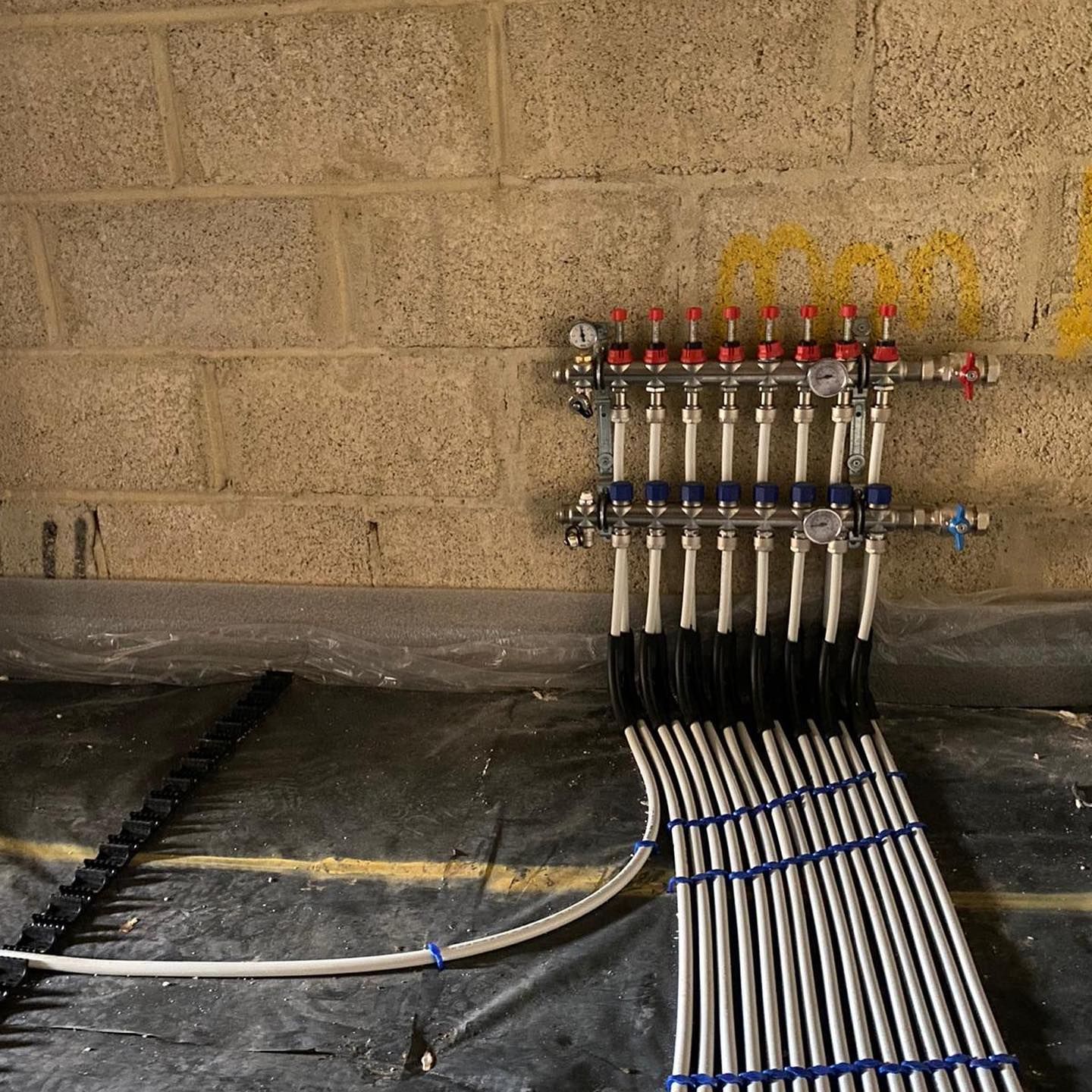 Radiant floor heating manifold with multiple supply and return pipes mounted on a cinder block wall.