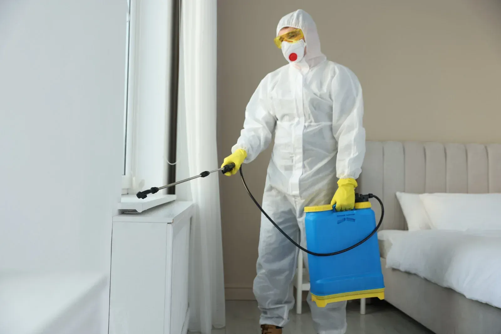 Person in protective suit spraying insecticide near a window in a bedroom.