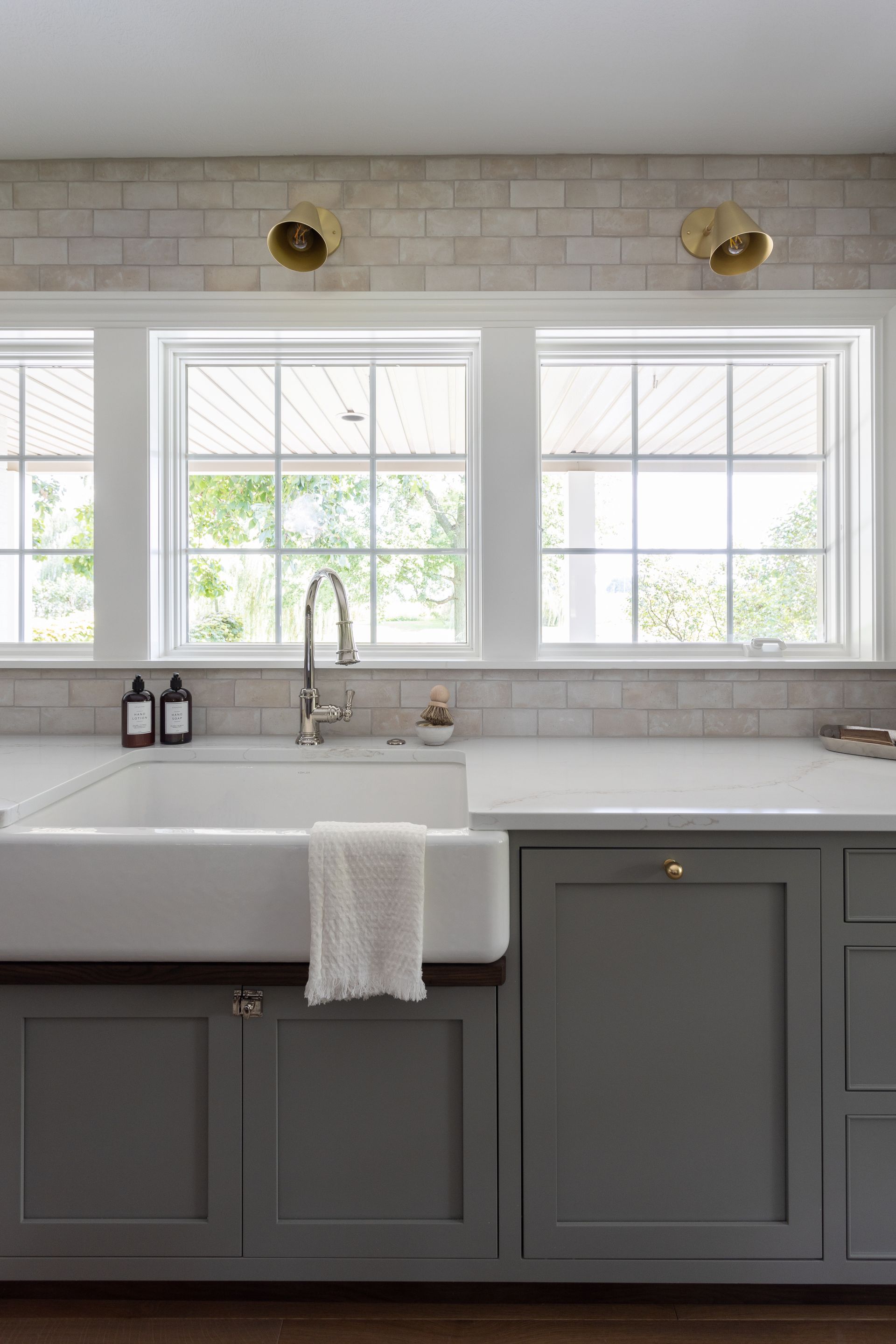 Kitchen with farmhouse sink, gray cabinets, white countertops, and three windows.