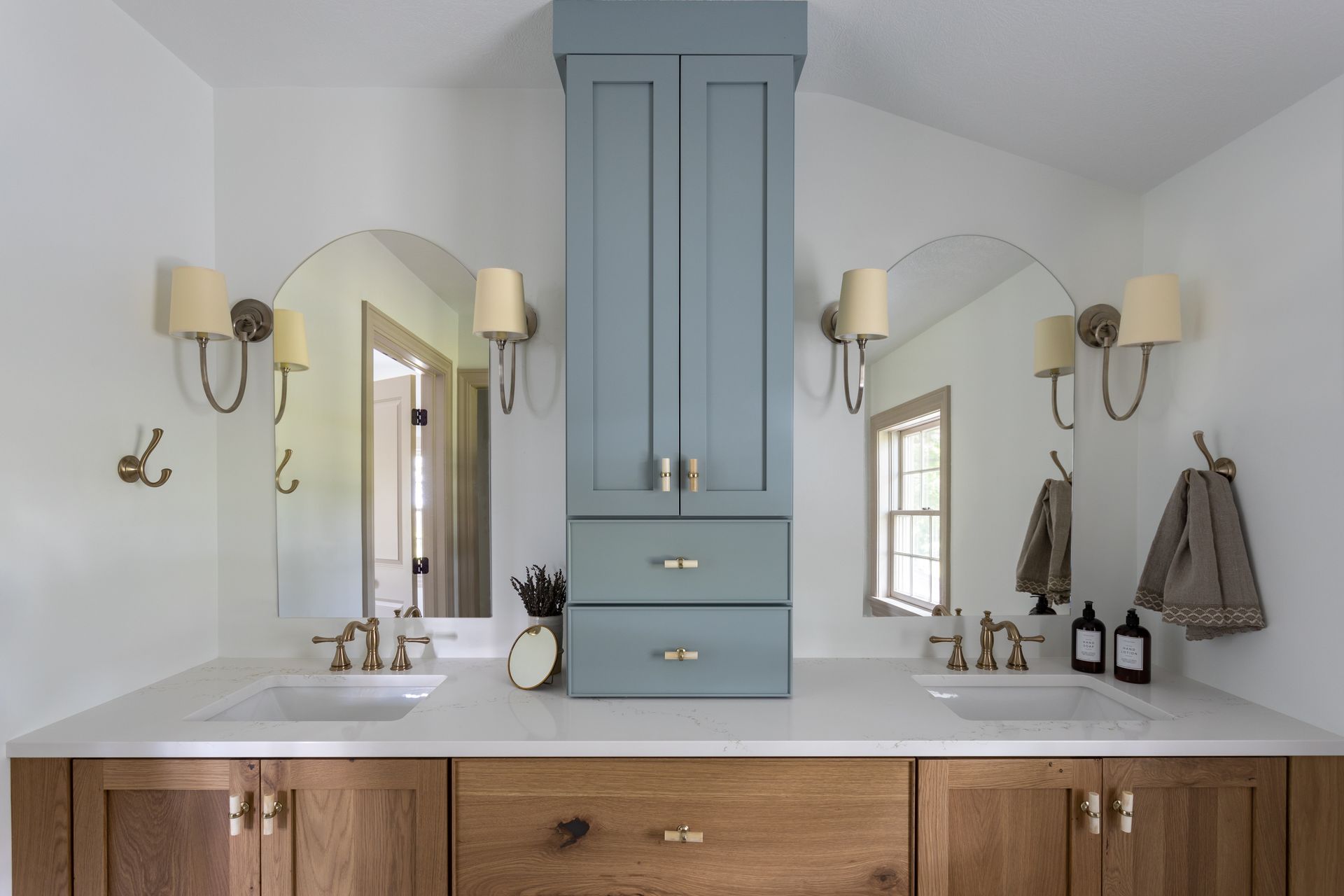 Bathroom with wooden vanity, white countertops, blue cabinet, arched mirrors, and sconces.